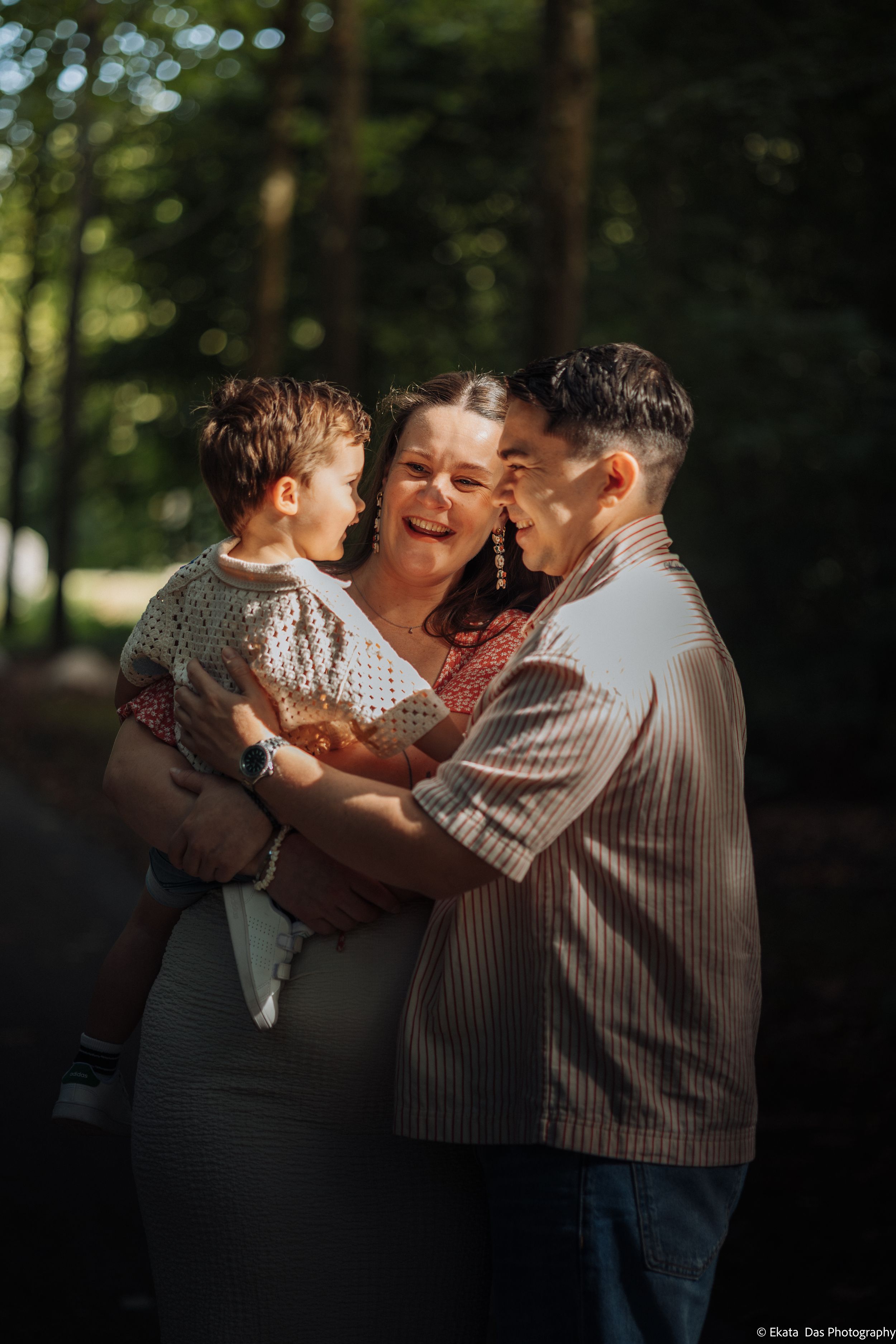 Smiling family of three embracing in a sunlit forest, a woman holding a toddler while a man hugs them close during an outdoor photo session.