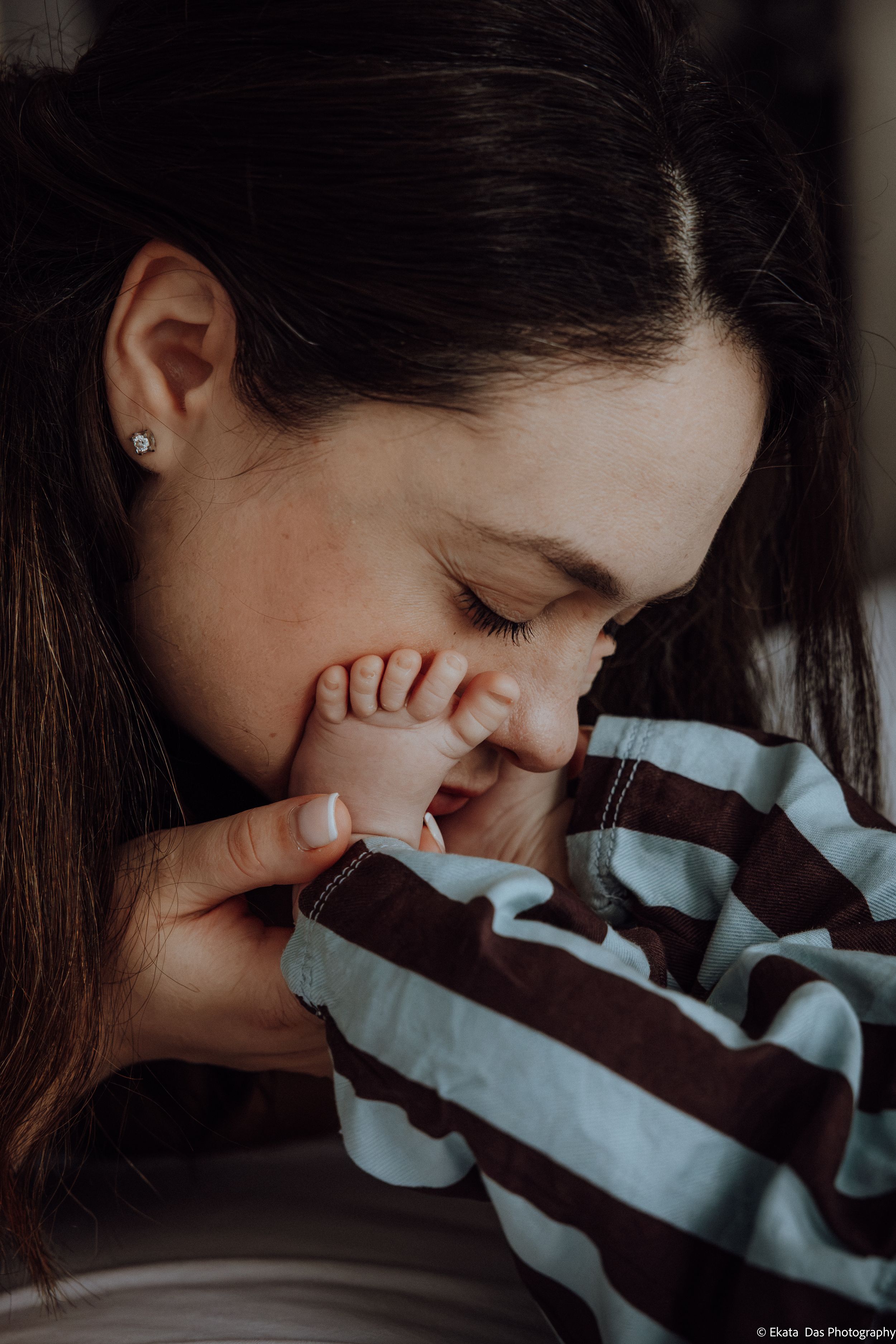 Mother in a striped sweater tenderly kisses her newborn baby's tiny foot in a warm, intimate lifestyle family portrait.