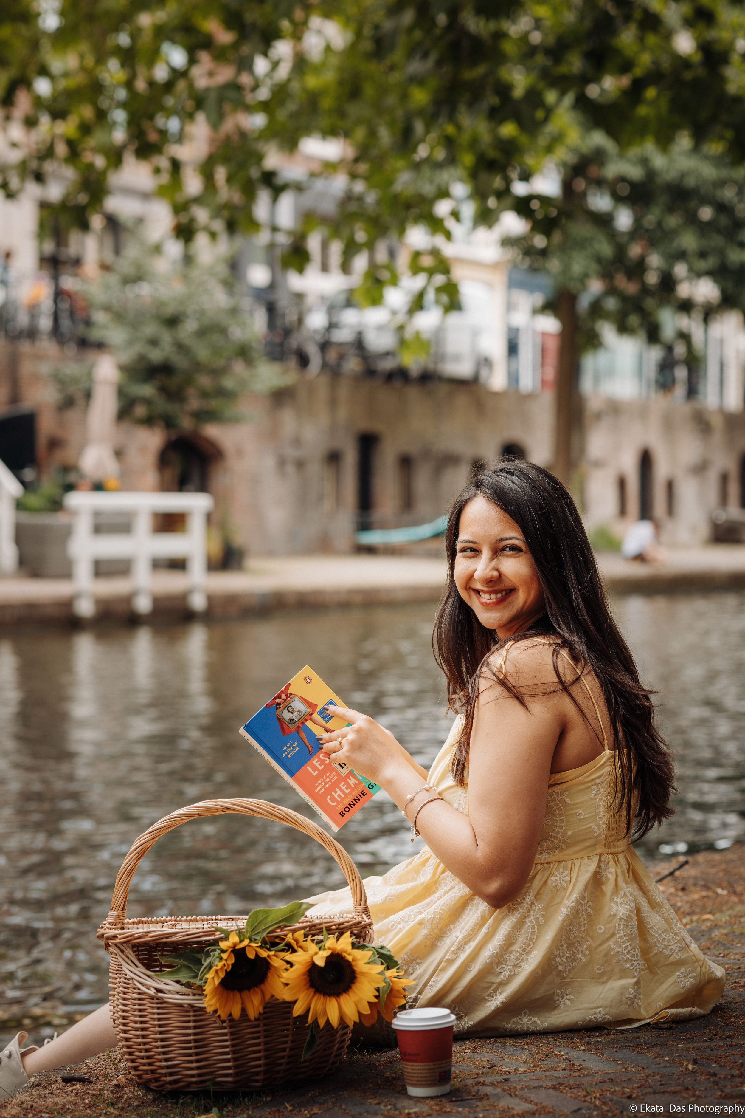 Smiling woman in a yellow sundress sits by a canal holding a book, with a wicker picnic basket, sunflowers and a coffee cup beside her.