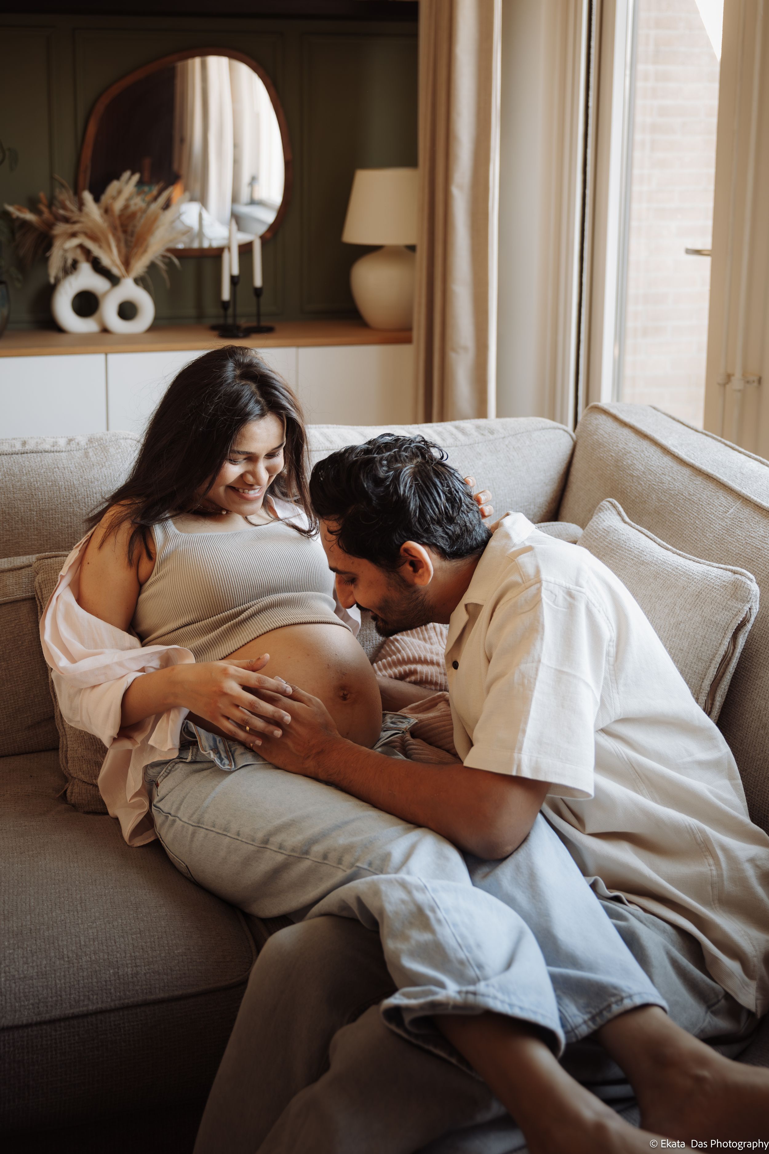 Expectant couple relaxing on a beige sofa as the smiling pregnant woman and her partner tenderly cradle her baby bump in a cozy living room.