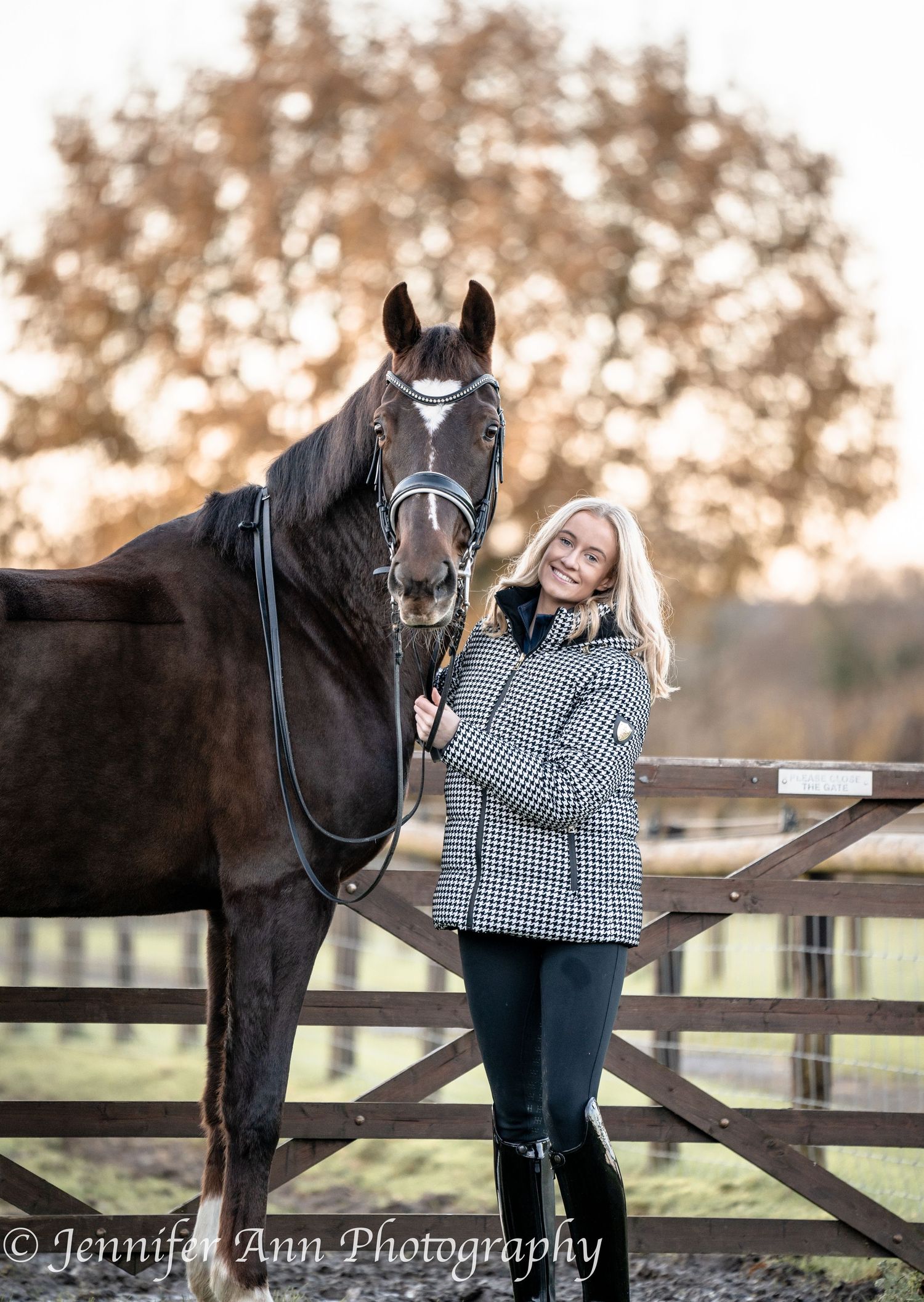 A beautiful black horse stands next to a wooden fence while wearing a bridle and being photographed outdoors.
