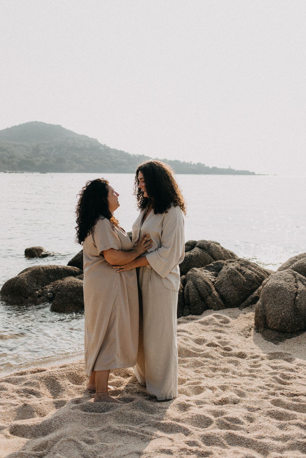 Two people in flowing beige dresses share an intimate moment on a rocky beach with mountains in the background.