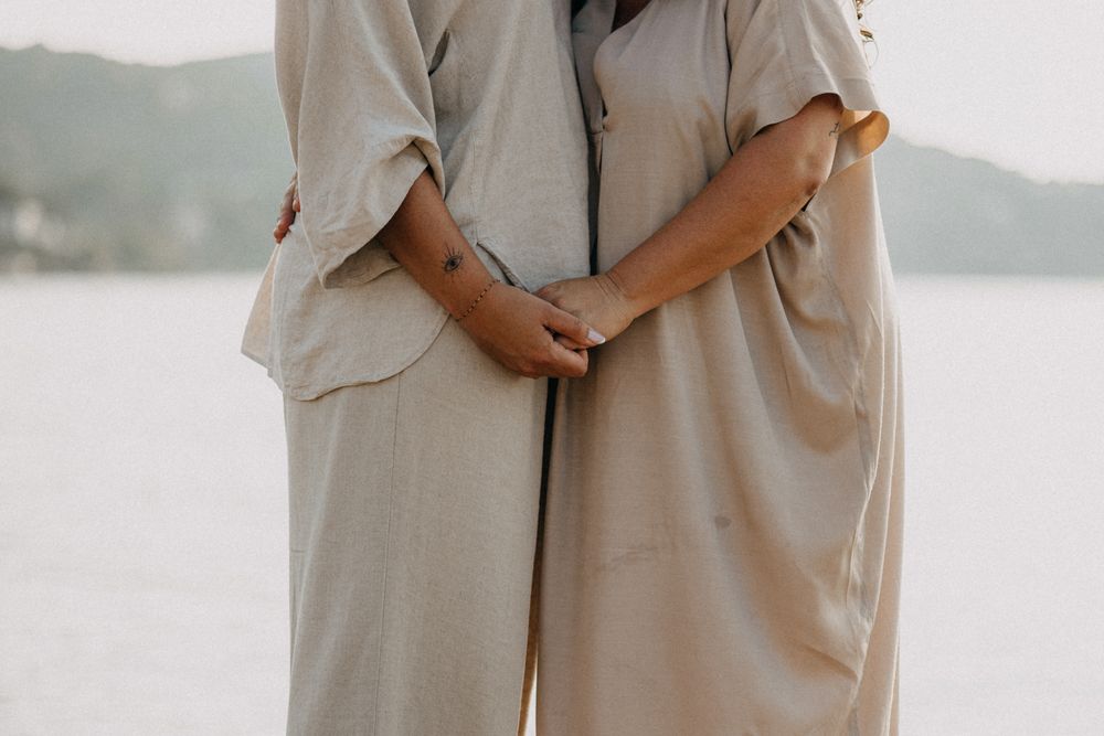 Two people in beige linen outfits hold hands while standing together by a serene waterfront.