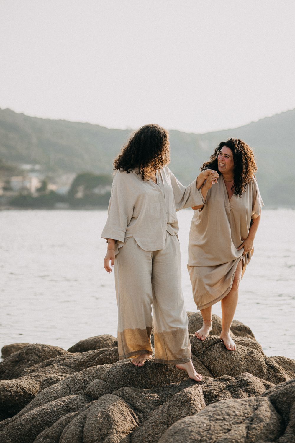Two people in beige outfits sit together on coastal rocks overlooking the ocean at sunset.