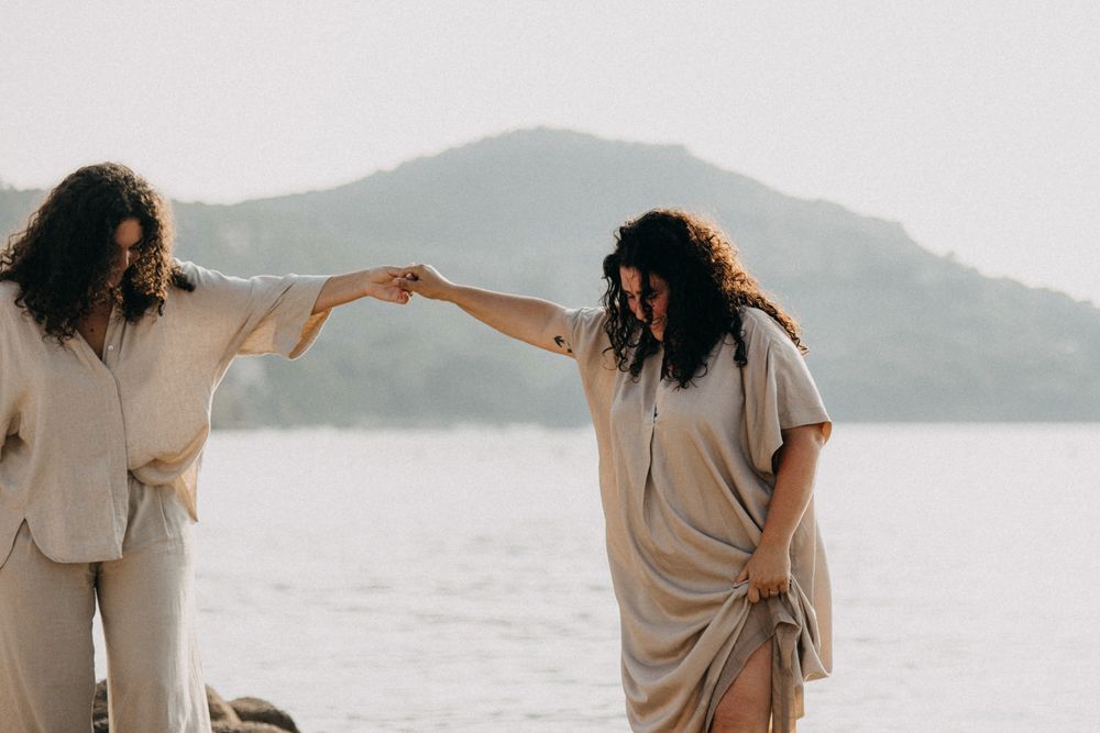 Two people in beige outfits dance together on a beach with mountains in the background at sunset.