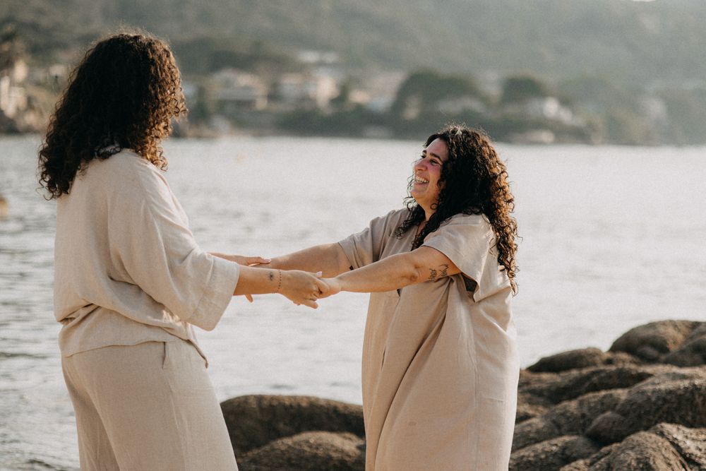 Two people in white outfits hold hands while standing on rocky beach during sunset.