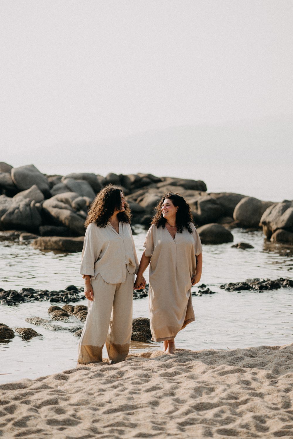 Two people in light colored outfits walking together on a rocky beach at sunset.