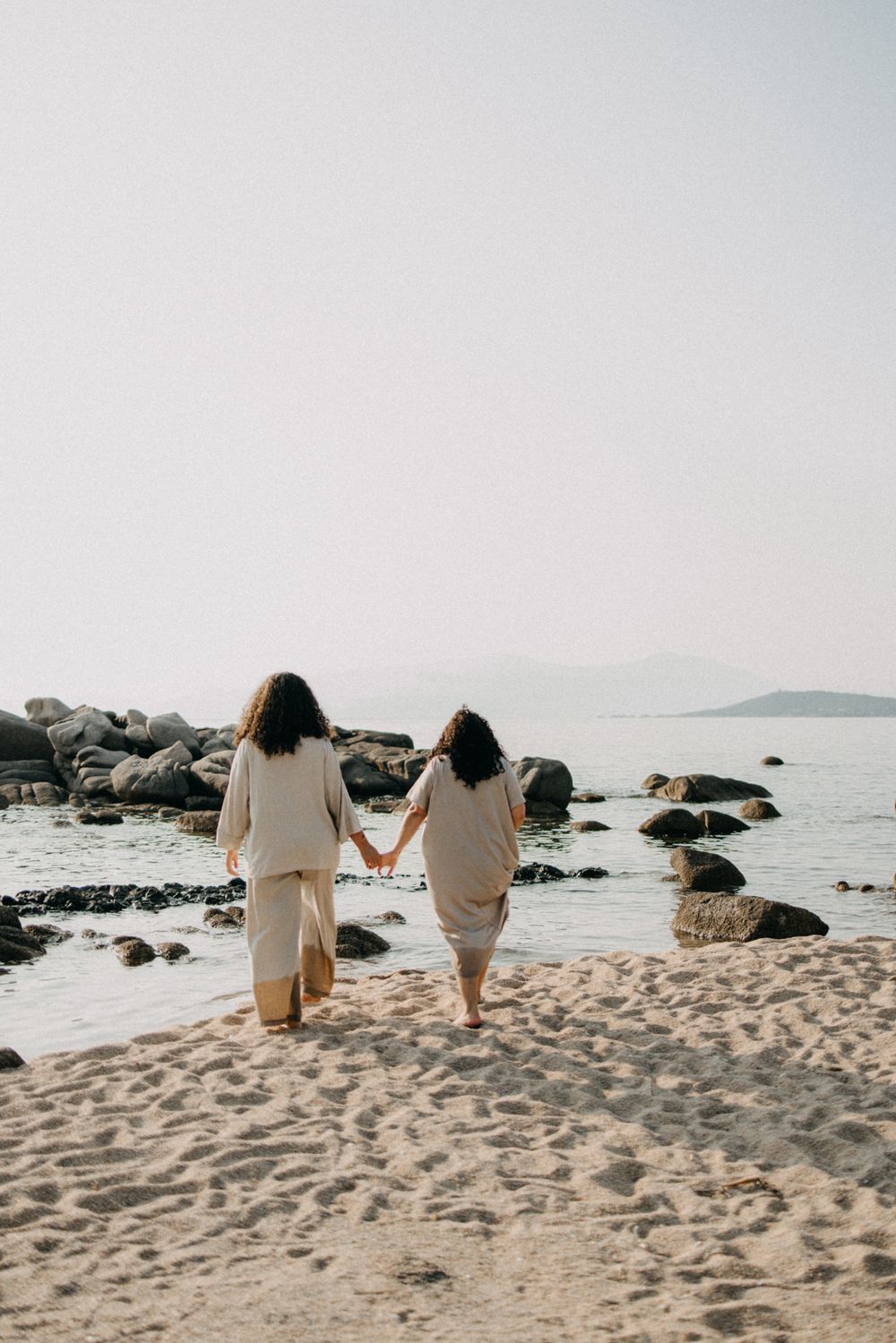 Two people in white outfits hold hands while walking on a rocky beach at sunset with ocean and mountains in view.