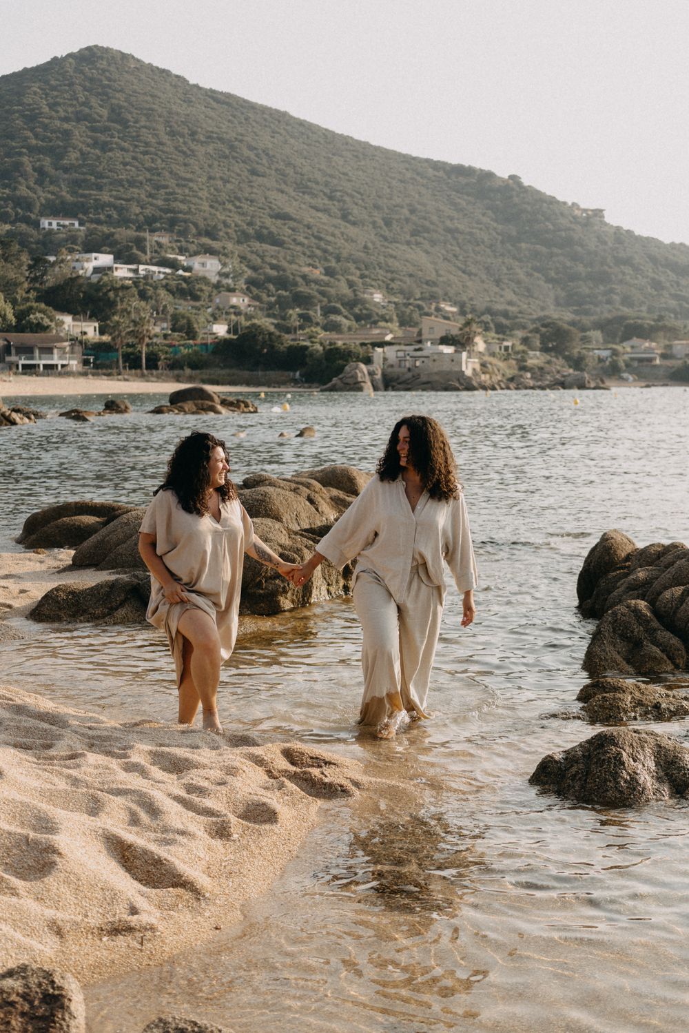 Two people in light colored clothing walking together along a rocky beach shore with mountains in the background.