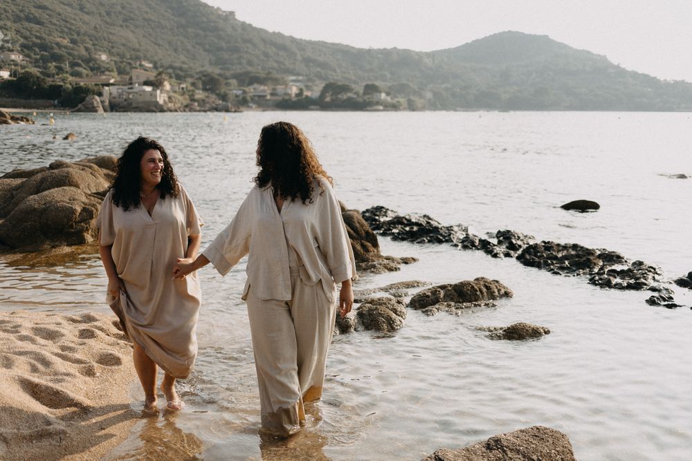 Two figures strolling hand in hand on a sandy beach with rocky coastline and hills in the distance.