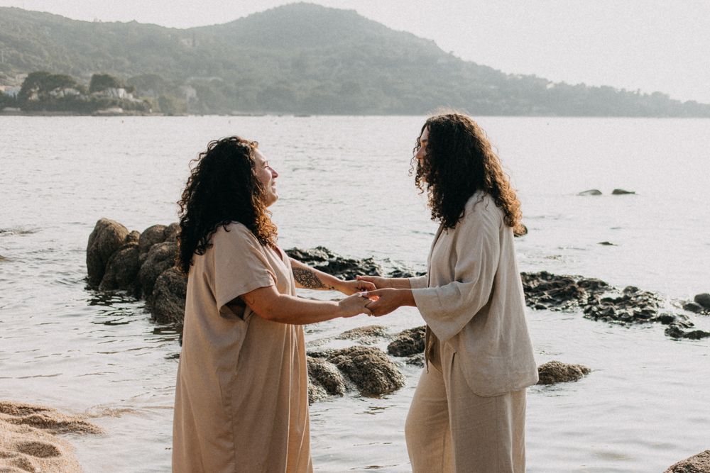 Two people in white outfits share intimate moments by the ocean with mountains in the background at sunset.