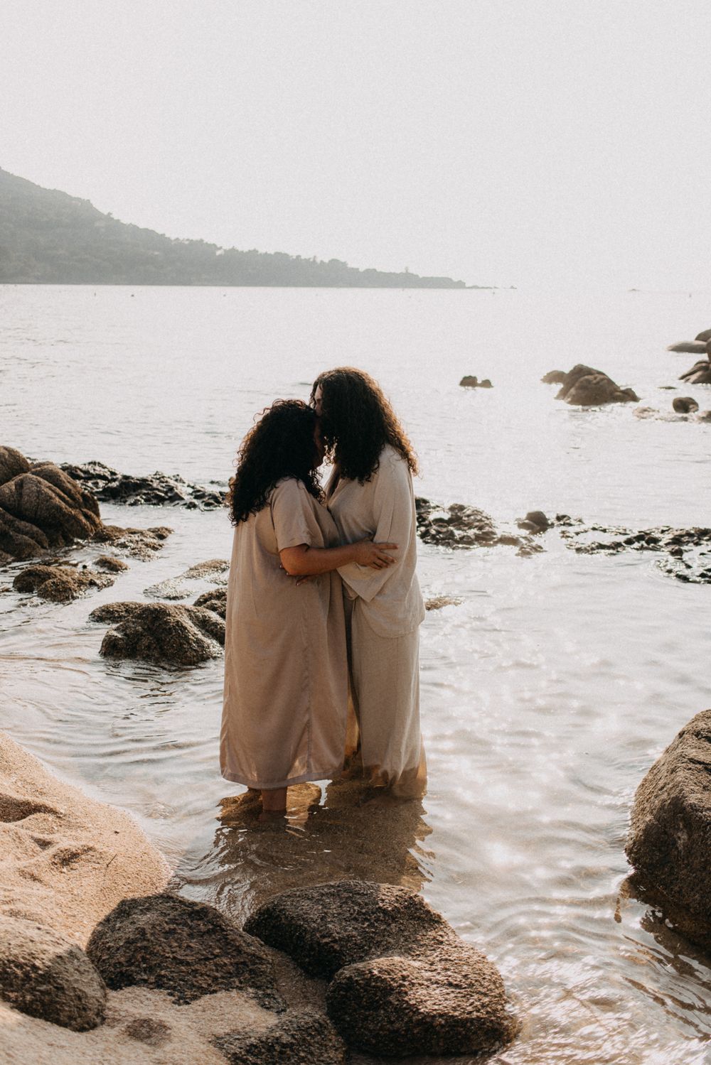 Couple sitting together on rocky beach shore watching ocean waves at sunset in matching beige outfits.