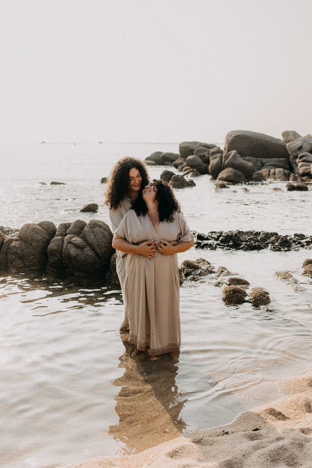 Two people embracing on a misty beach surrounded by large rocks and gentle waves.