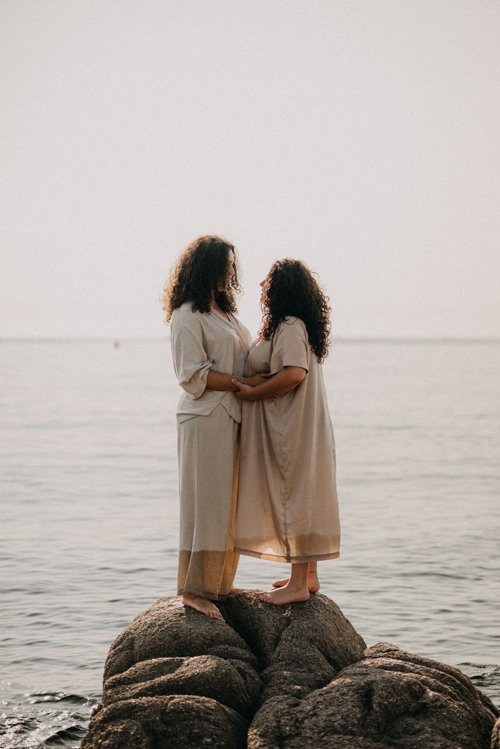 Two figures in white dresses stand together on coastal rocks overlooking a serene ocean view at sunset.