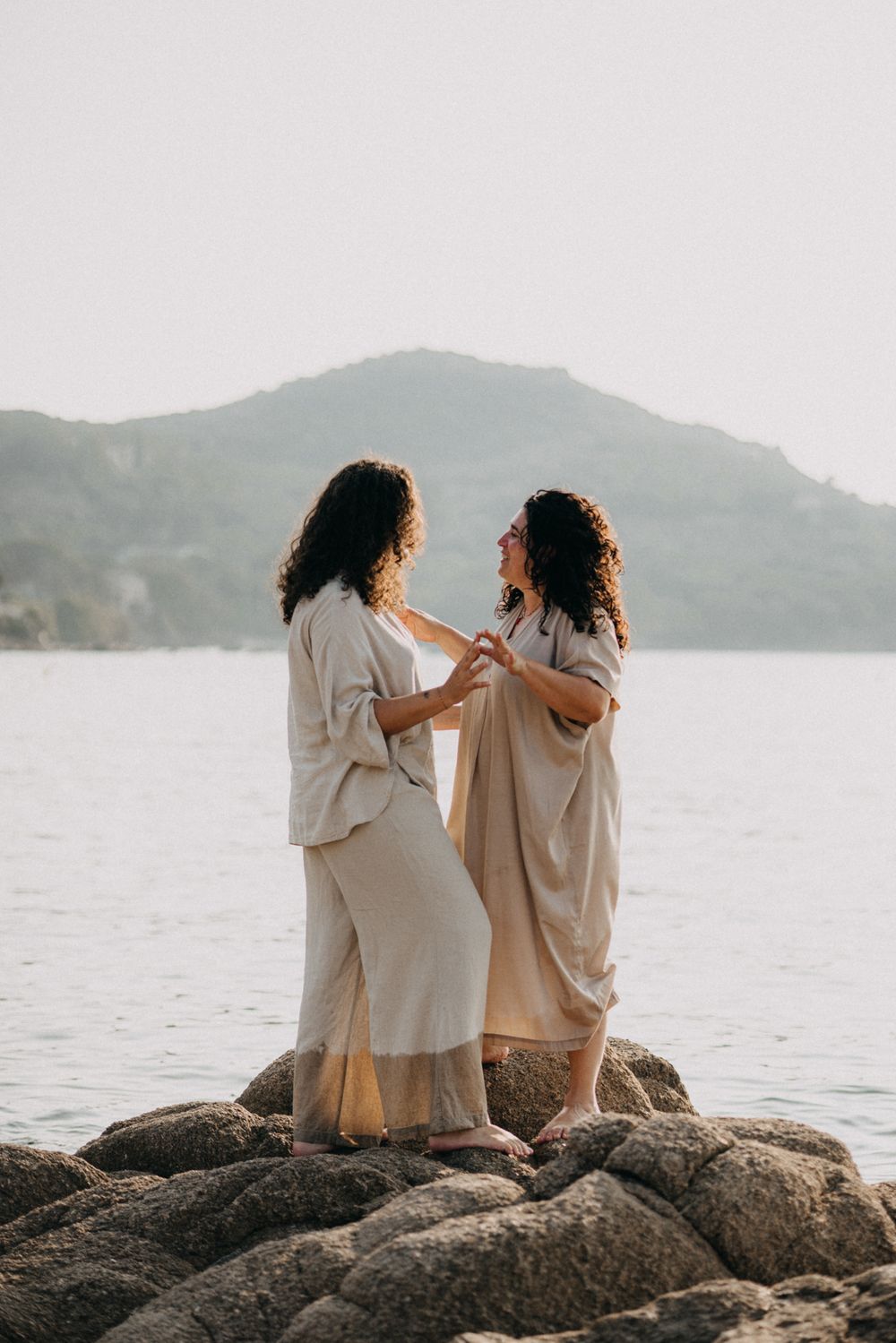 Two people in light colored clothing sit together on coastal rocks at sunset with mountains in the background.