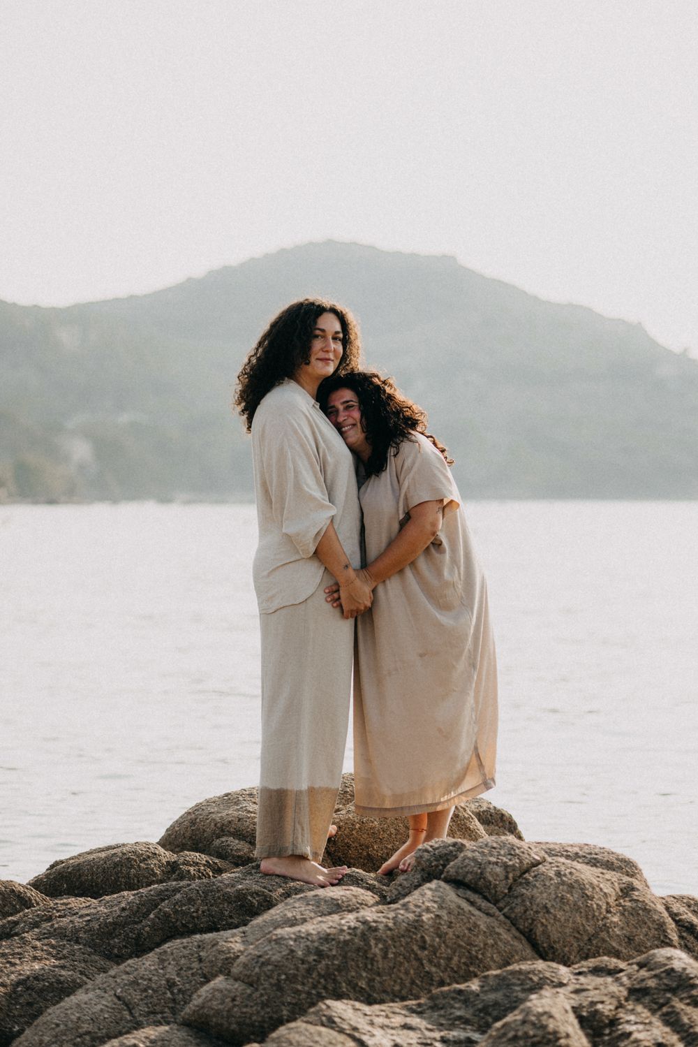 A couple in white clothing embraces while sitting on rocky coastal outcrop with mountains and water in background.