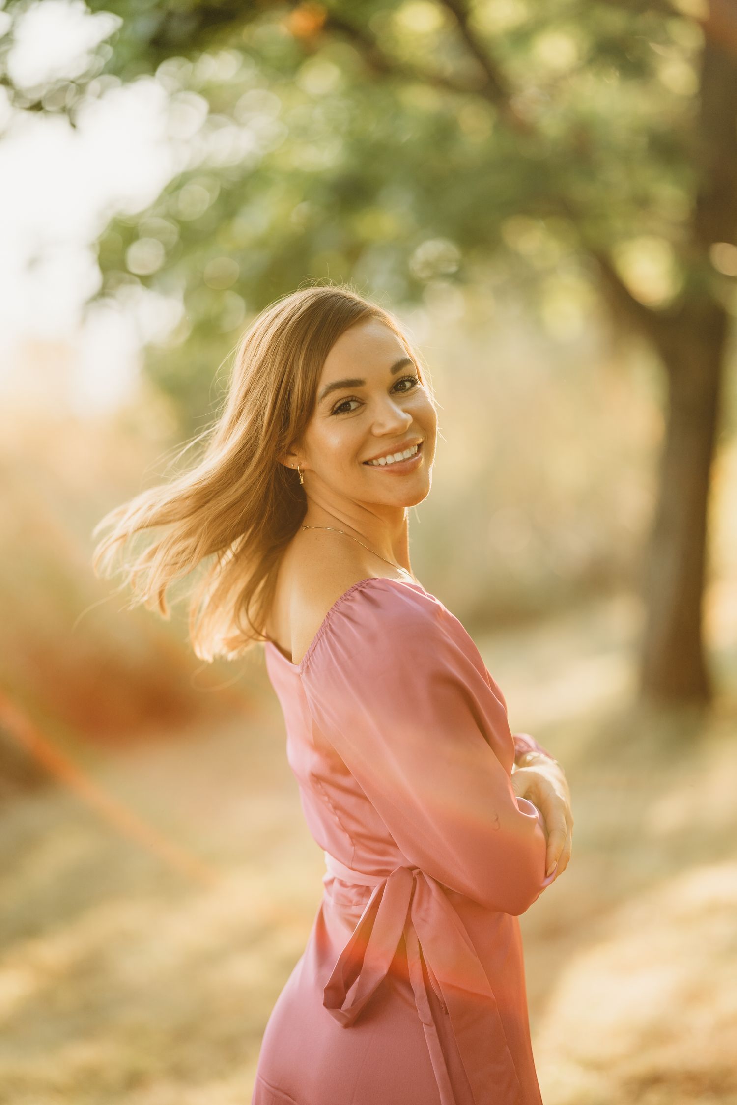 A person in a pink dress smiles while standing outdoors in warm sunlight with a blurred natural background.
