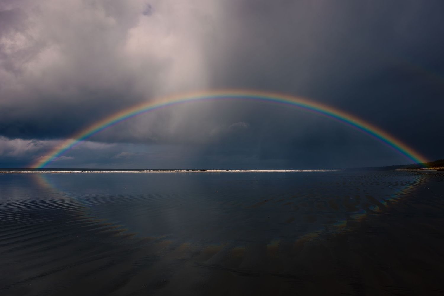 A vibrant double rainbow arches over a dark ocean horizon with dramatic storm clouds in the background.