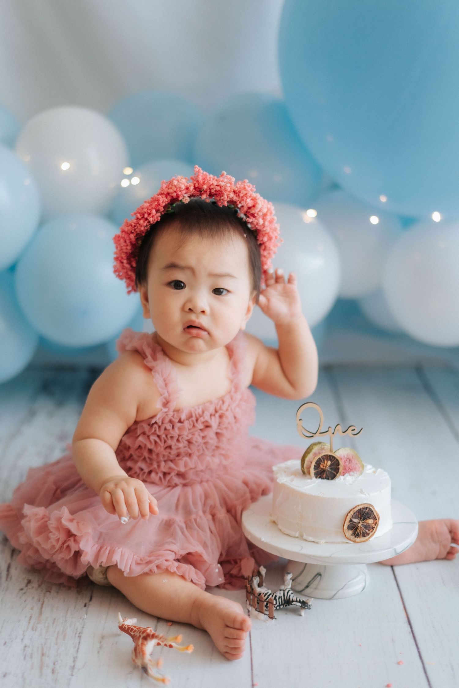 A baby sits in a white chair surrounded by floral decorations and balloons at a birthday celebration.