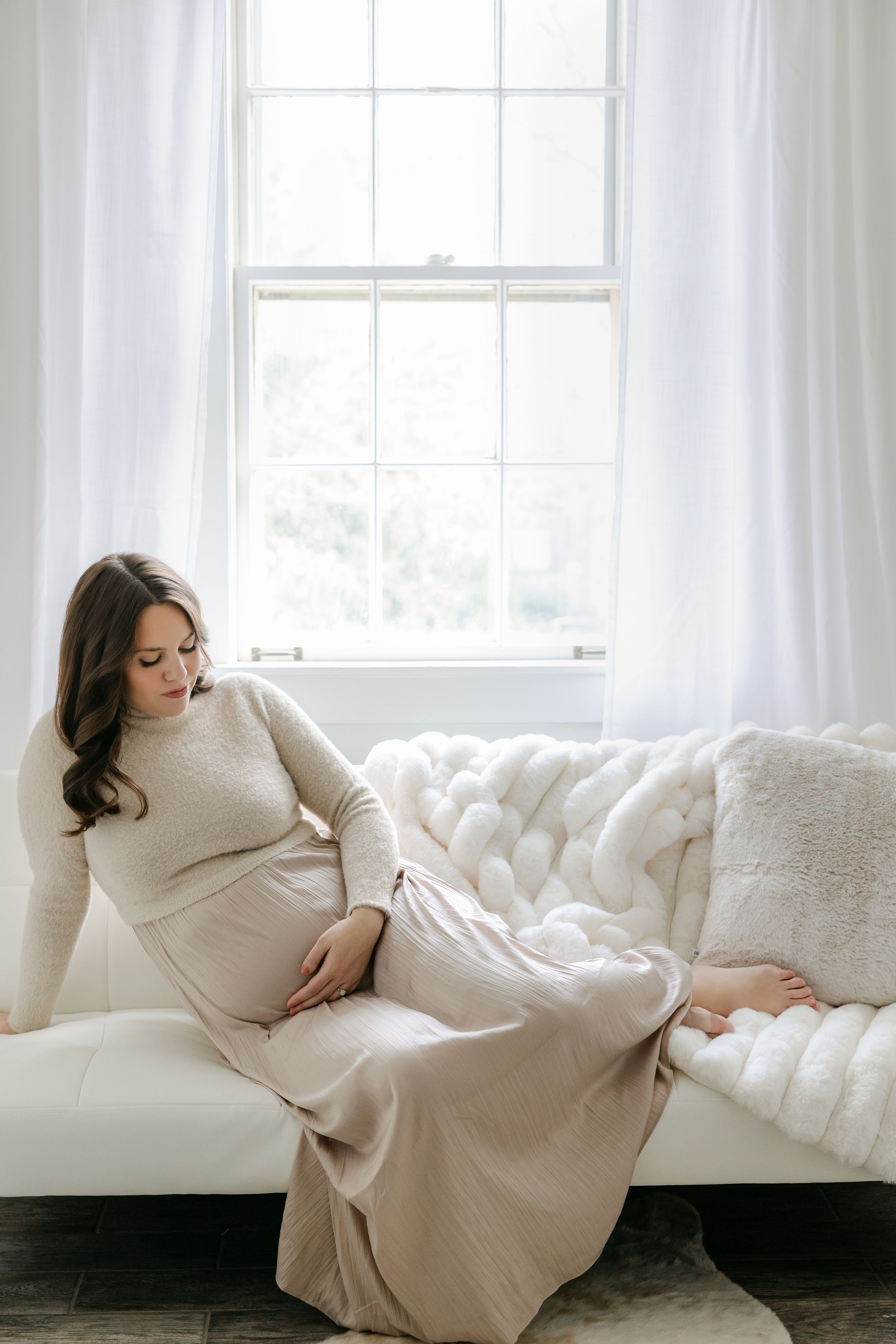 A woman in a cream sweater dress reclines gracefully on a white tufted sofa near a bright window.