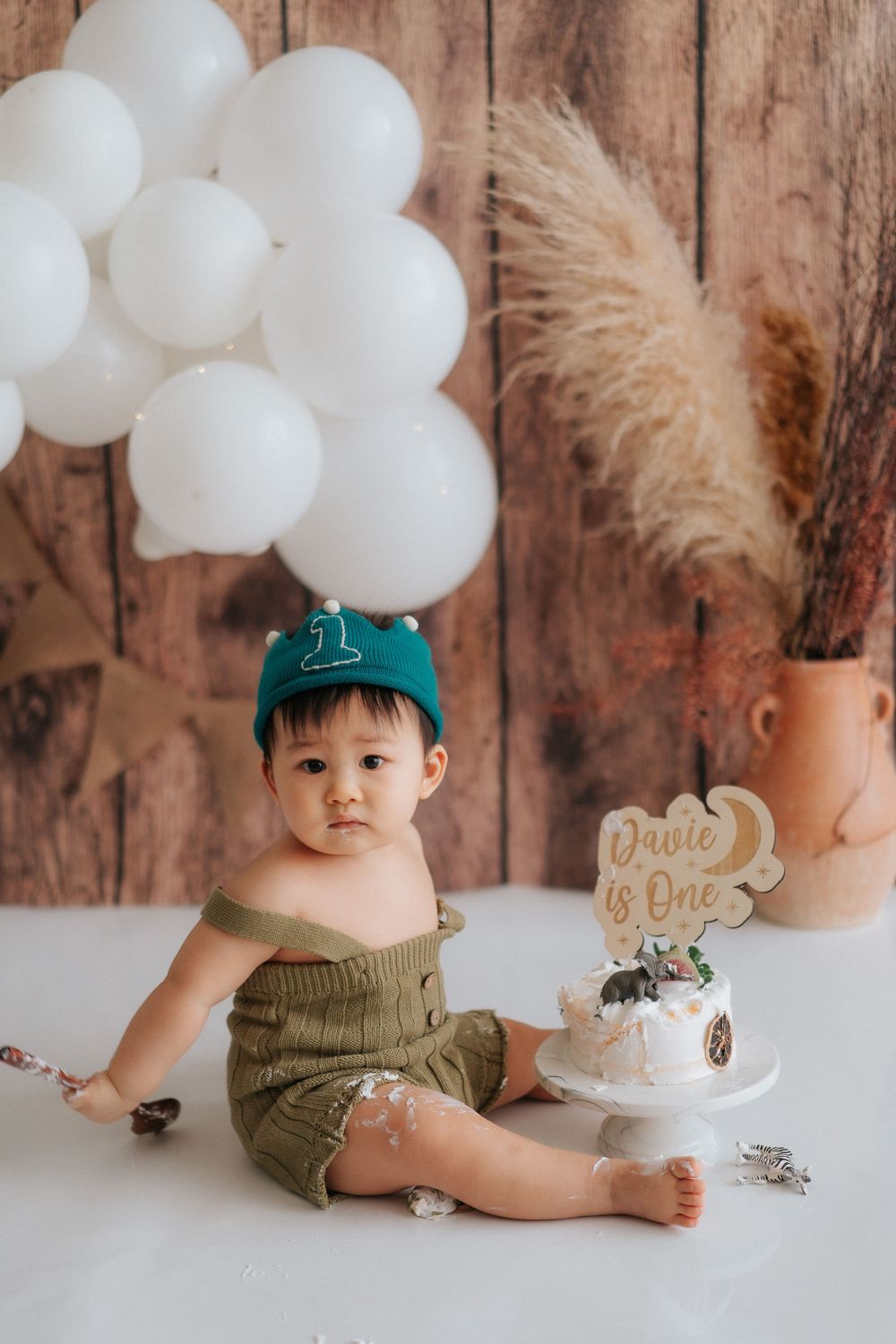 First birthday celebration setup with white balloons, cake, and pampas grass against rustic wooden backdrop.