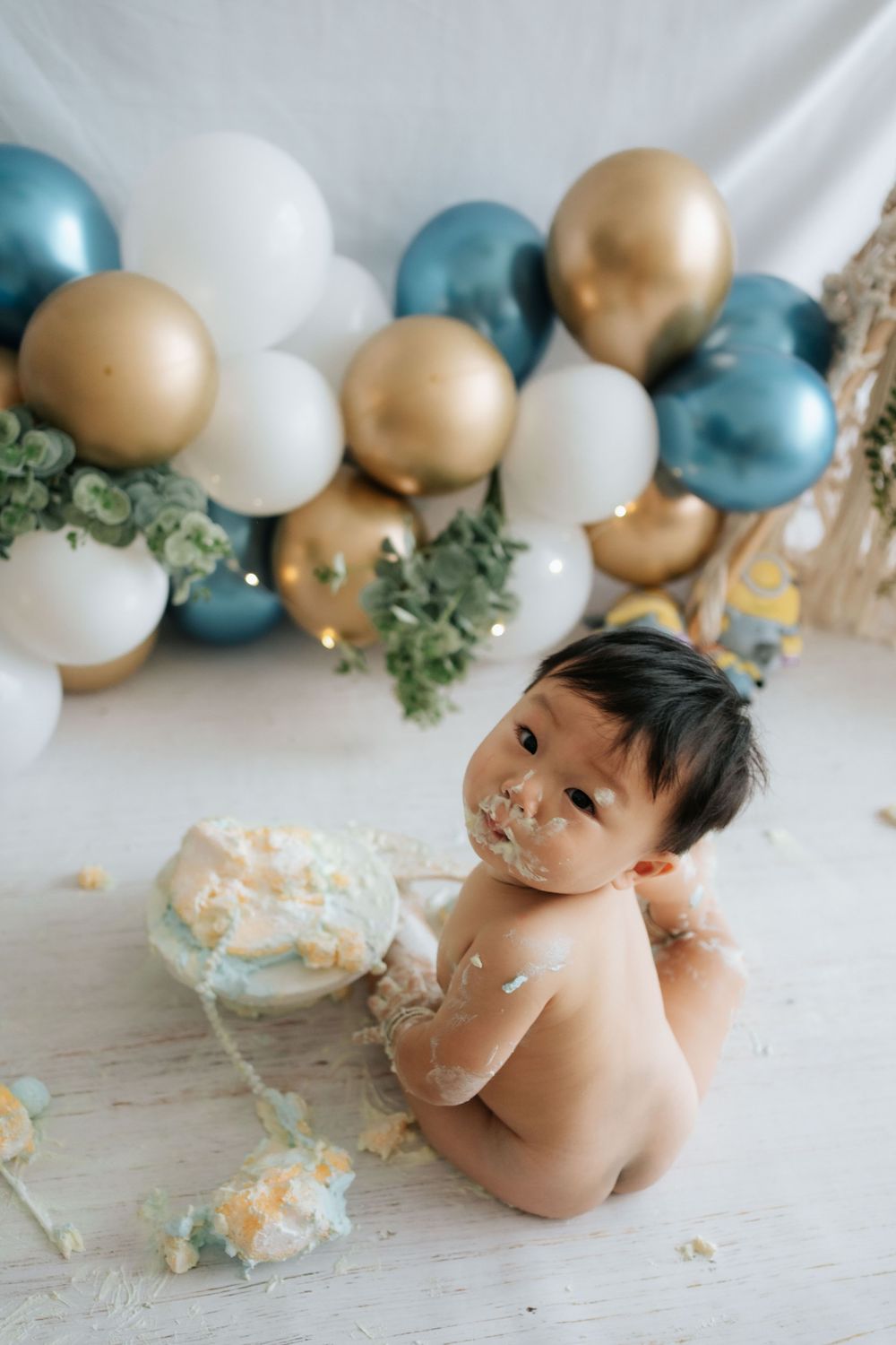 Baby cake smash photo session with teal and gold balloon arch backdrop and scattered cake pieces.