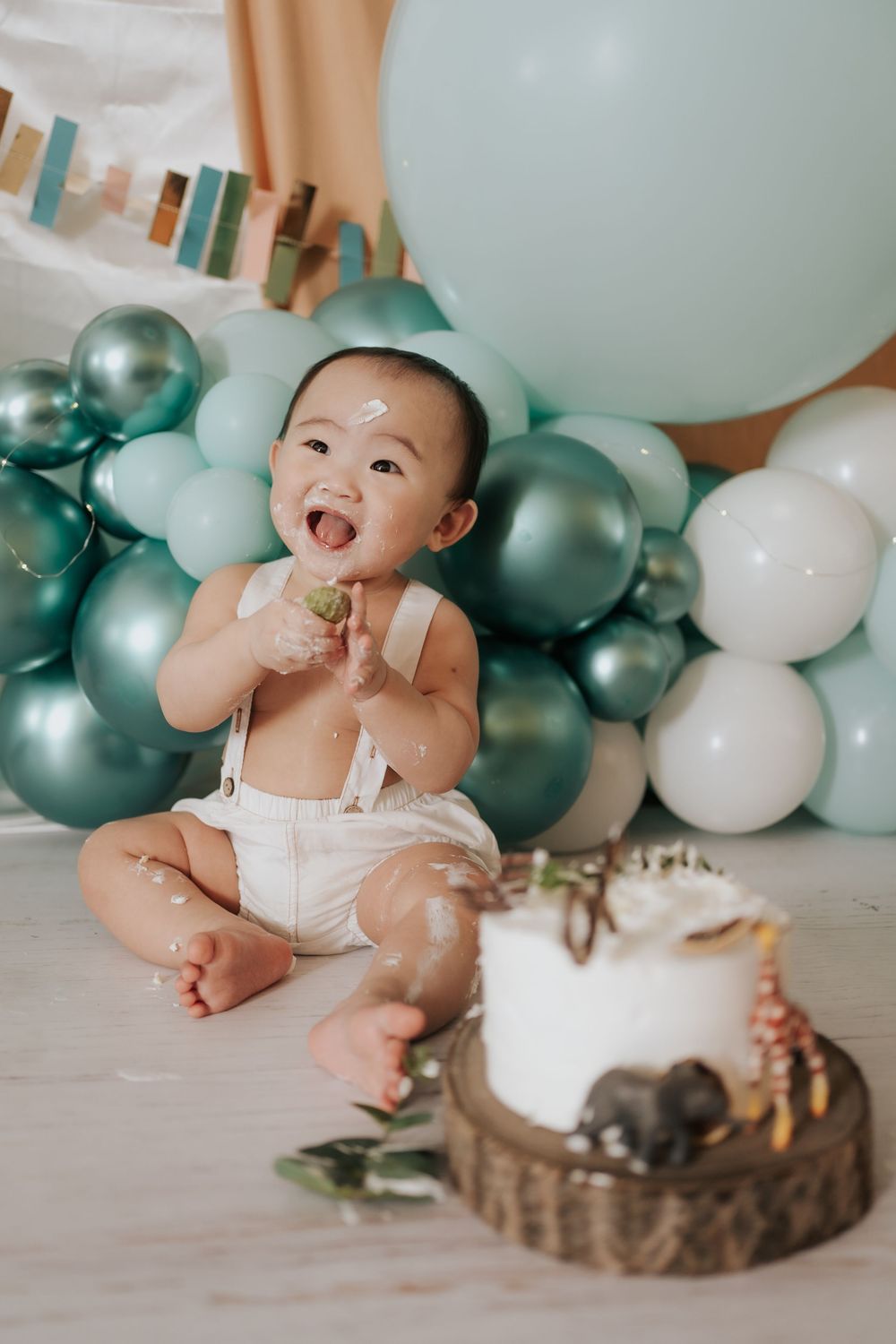 A baby in white clothing sits on the floor for a cake smash photo session with teal and white balloons in the background.