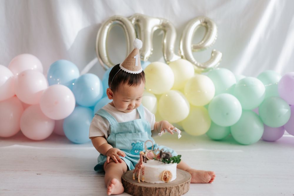 A baby in a white outfit sits on the floor with pastel balloon decorations and a birthday cake for their first birthday celebration.