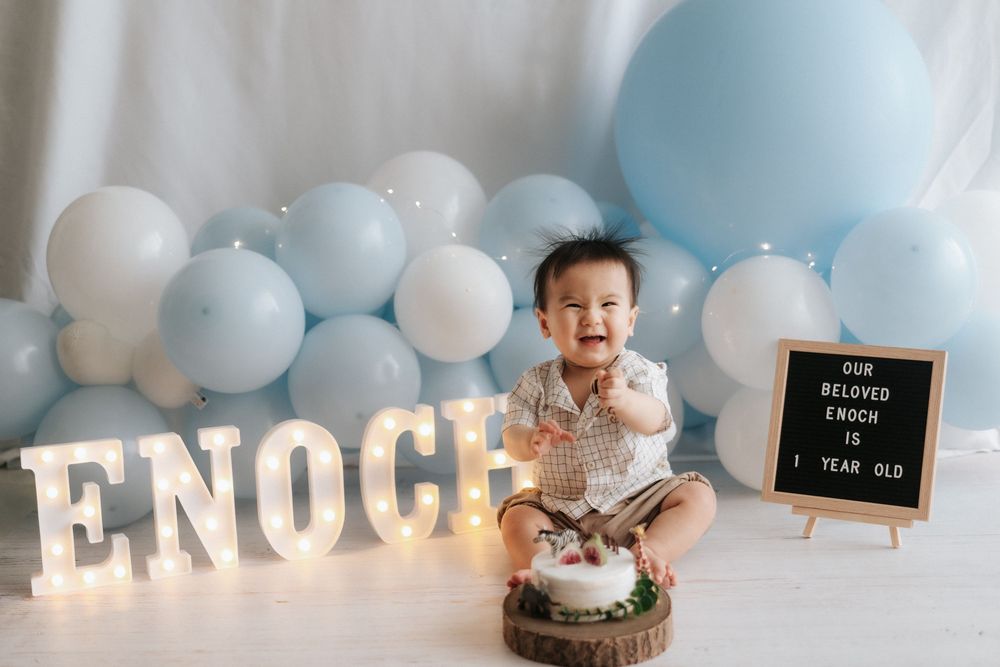 First birthday celebration setup with light-up letters, blue and white balloons, and wooden decor against light background.