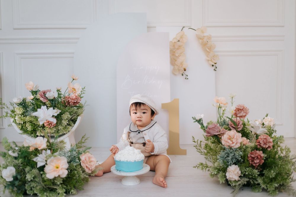 A series of cake smash photos showing a baby in white celebrating their first birthday with a blue cake and floral decor.