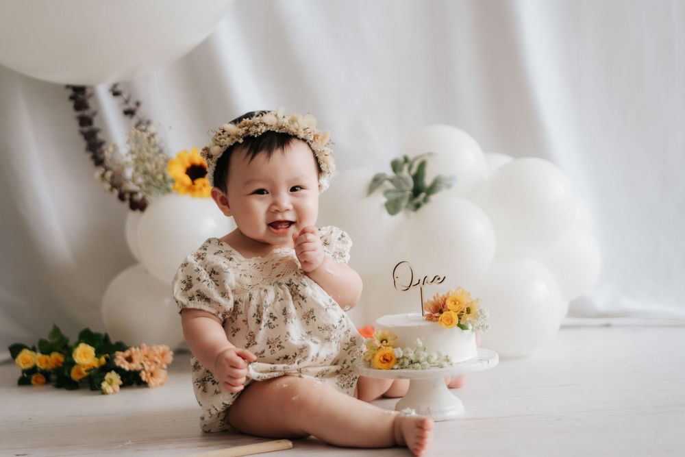 A baby in a white lace dress sits on a white blanket surrounded by sunflowers and autumn decor.
