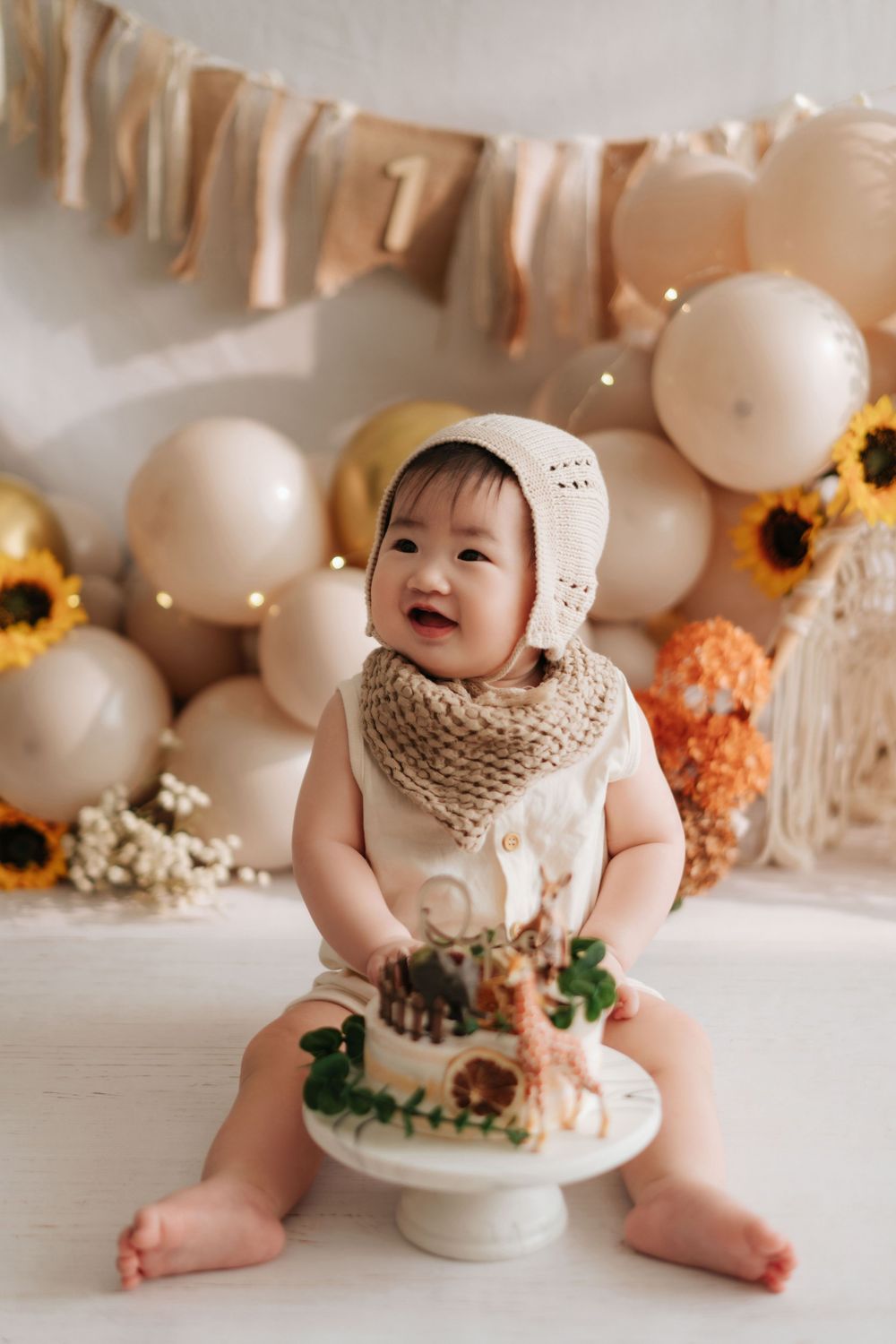 A cake smash photo session featuring a baby with white balloons and sunflower decorations in warm neutral tones.