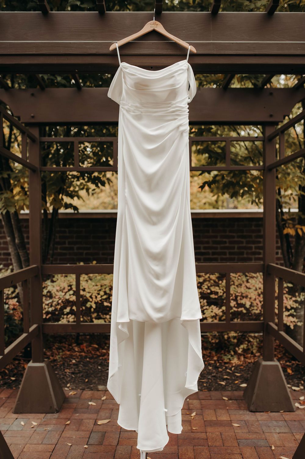 Elegant white satin wedding gown hanging on wooden hanger in outdoor pergola with autumn foliage.