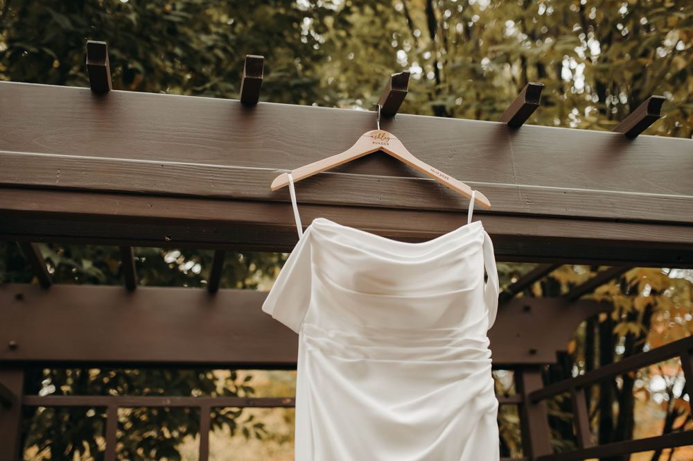 Close-up of white satin bridal gown with draped neckline hanging on wooden hanger outdoors.