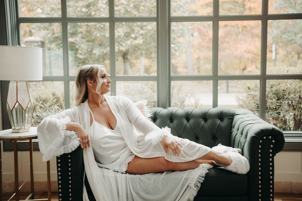 Bride in white robe relaxing on dark green tufted sofa by large windows with autumn view.