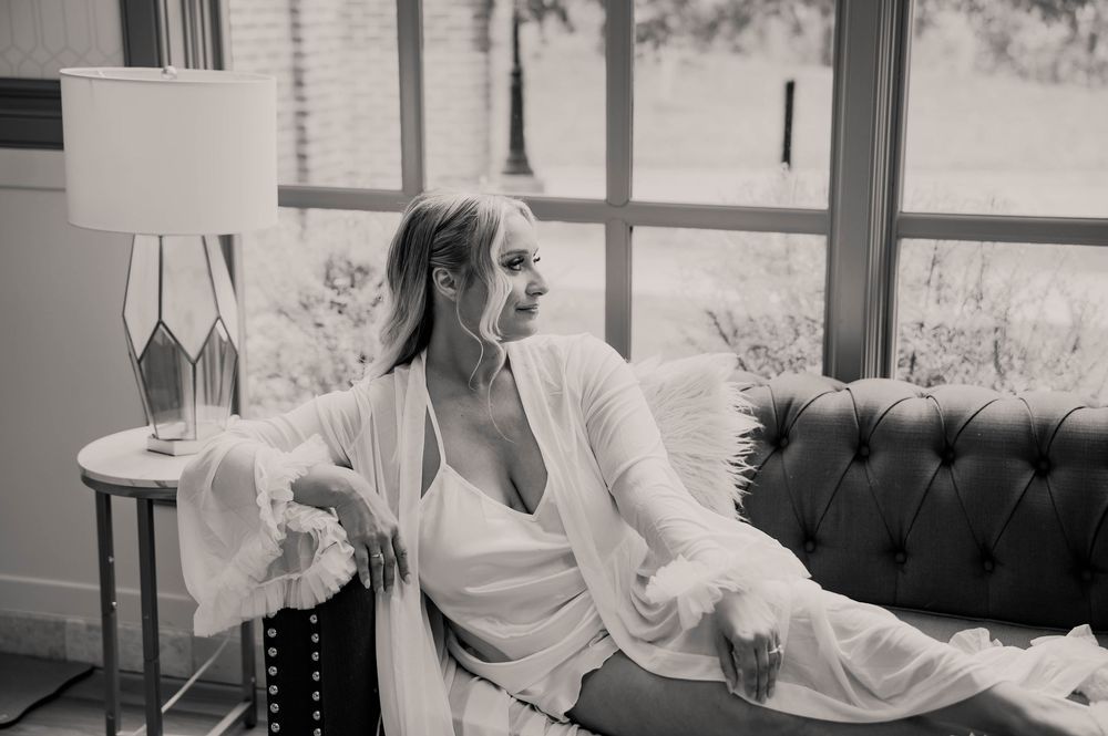 Black and white photo of bride in white robe gazing out large windows while seated on sofa.