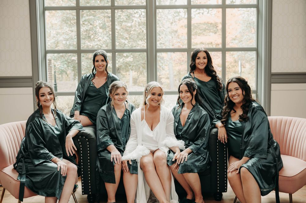 Bride in white robe posing with six bridesmaids in dark green satin robes on pink chairs.