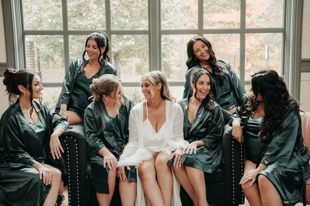 Bride surrounded by laughing bridesmaids in dark green satin robes near bright venue windows.