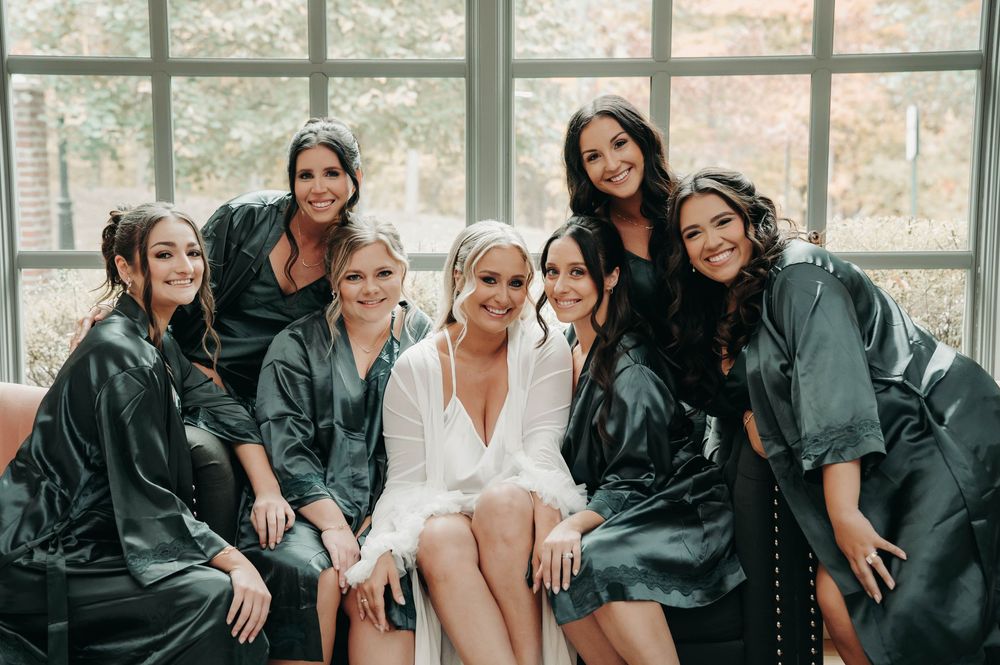 Bride in white feather robe smiling with six bridesmaids in dark green robes by window.