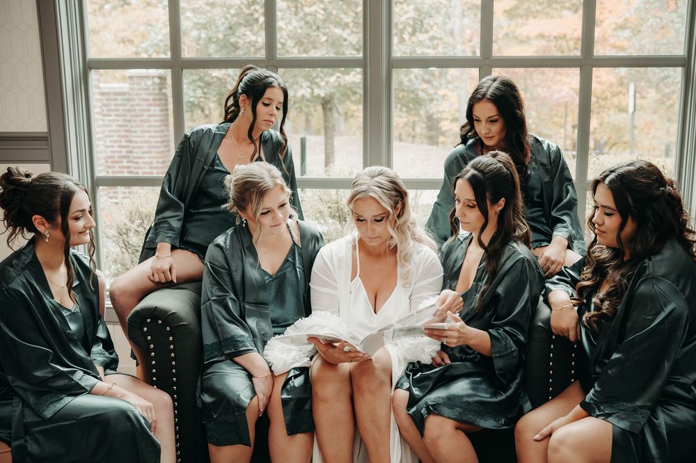 Bride reading a letter surrounded by attentive bridesmaids in dark satin robes by window.