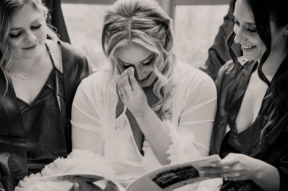 Emotional bride wiping tears while reading a letter flanked by two bridesmaids, black and white.