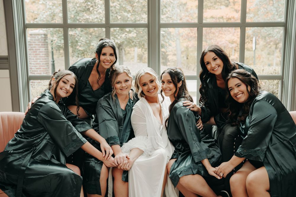 Bride in white robe poses with six bridesmaids in dark green satin robes before the wedding ceremony.