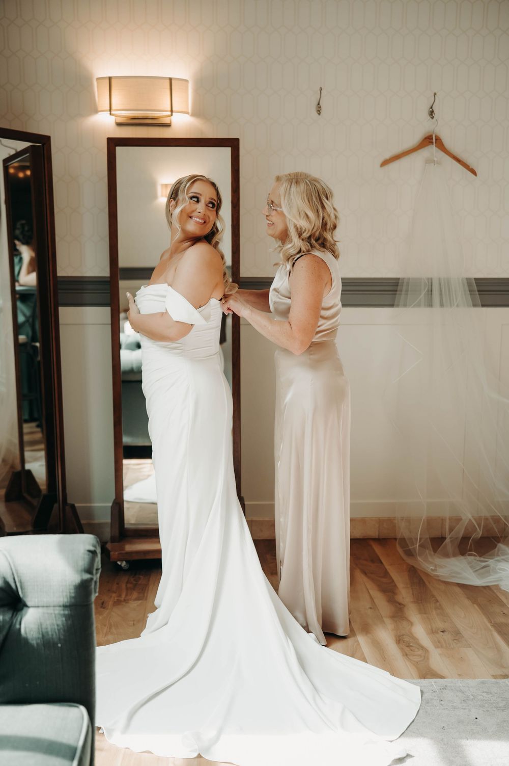Smiling bride in off-shoulder white gown gets dressed with help from mother in champagne satin dress.