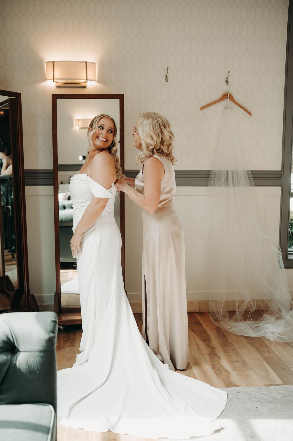 Bride in fitted white wedding dress smiles as mother buttons her gown near a full-length mirror.