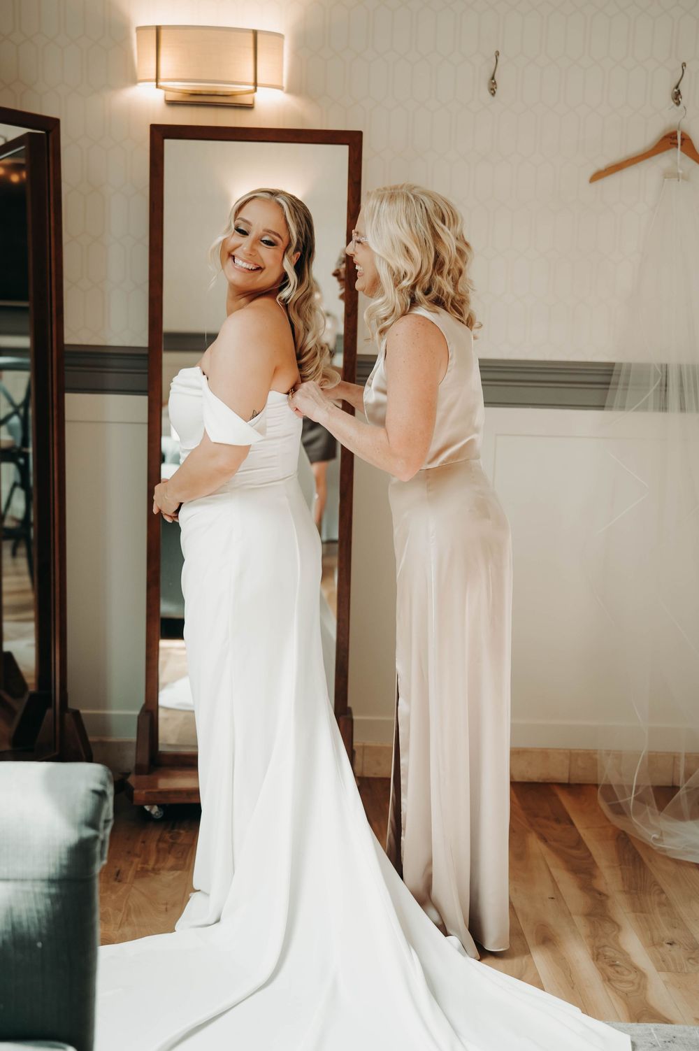 Radiant bride in off-shoulder white gown laughs while mother adjusts her dress before the ceremony.