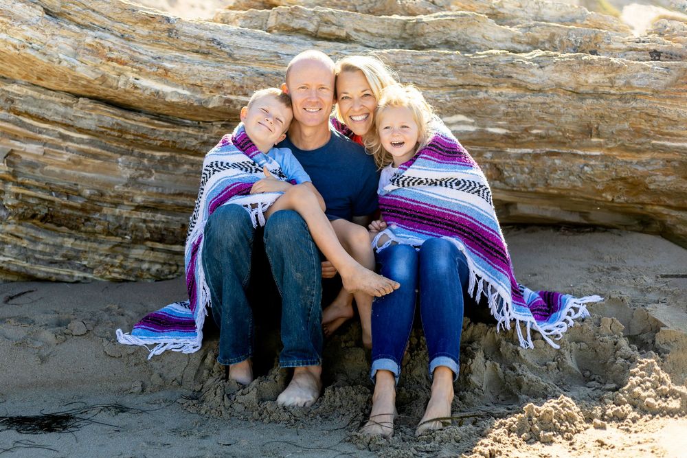 A group sits together wrapped in a purple striped blanket against a large piece of driftwood at the beach.