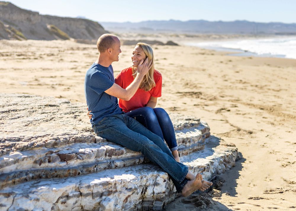 A couple sits together on driftwood at a scenic beach with mountains in the background on a sunny day.