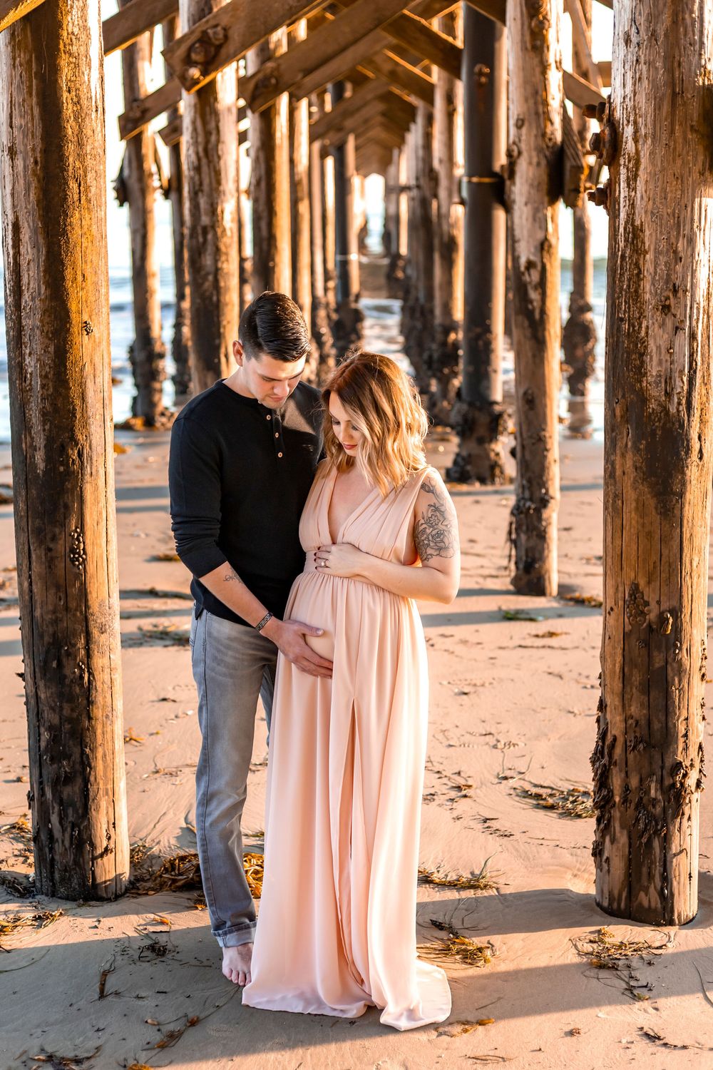 Romantic couple shares tender moments beneath a wooden pier during golden hour at the beach.
