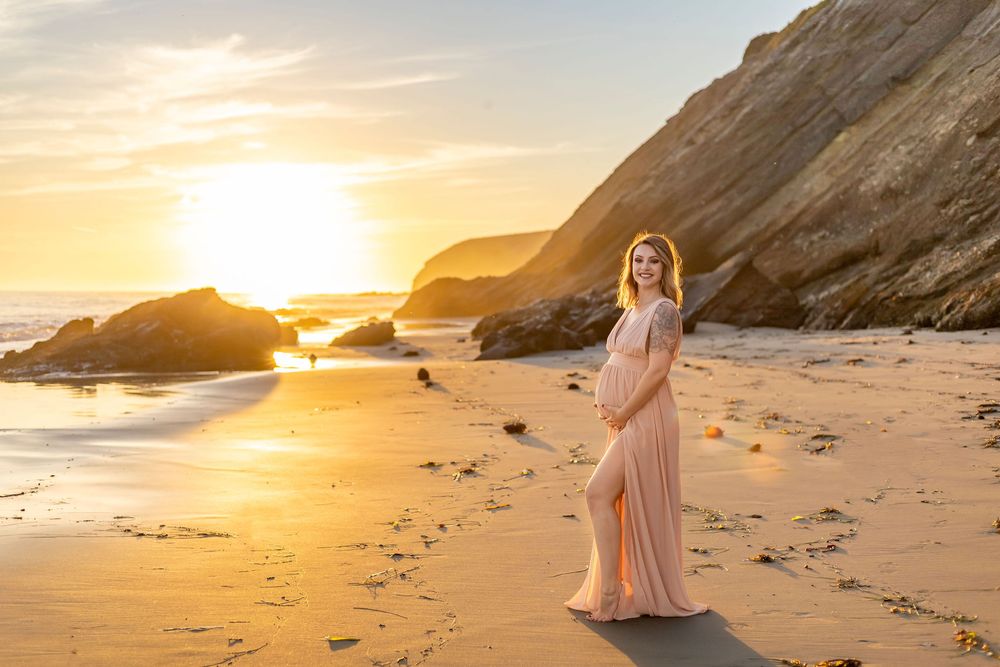 A scenic beach at sunset with golden light illuminating the shoreline and cliffs along the coast.  A pregnant woman in long pink dress