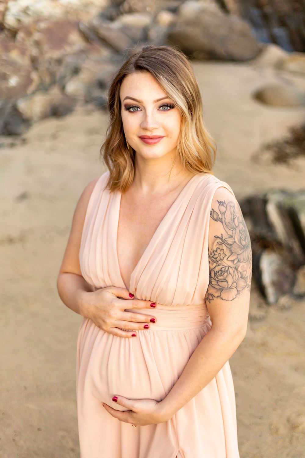A pregnant woman in a blush pink dress poses outdoors against a rocky backdrop in natural lighting on the beach